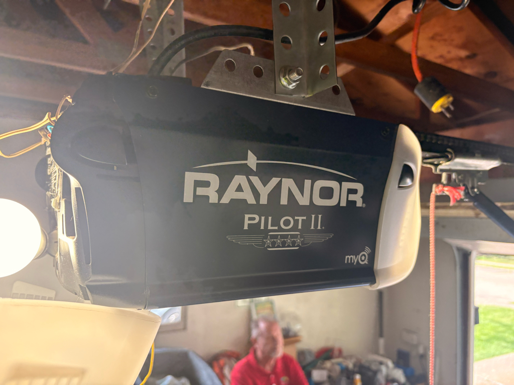 Raynor Pilot II myQ garage door opener mounted to a garage ceiling, with exposed wiring, hardware, sunlight, and a person in the background.