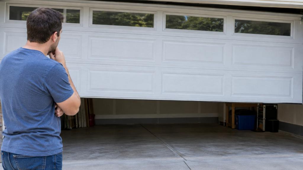 A concerned man watches a white garage door lift, revealing an empty, dimly lit garage with storage bins and concrete flooring.