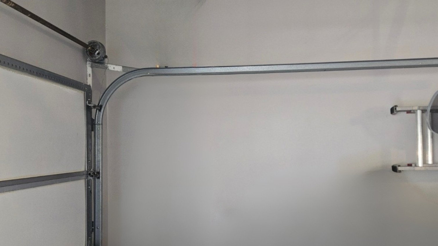 White sectional garage door seen from inside, with curved metal tracks, hinges, and support hardware along the upper-left corner; the scene feels plain and utilitarian.